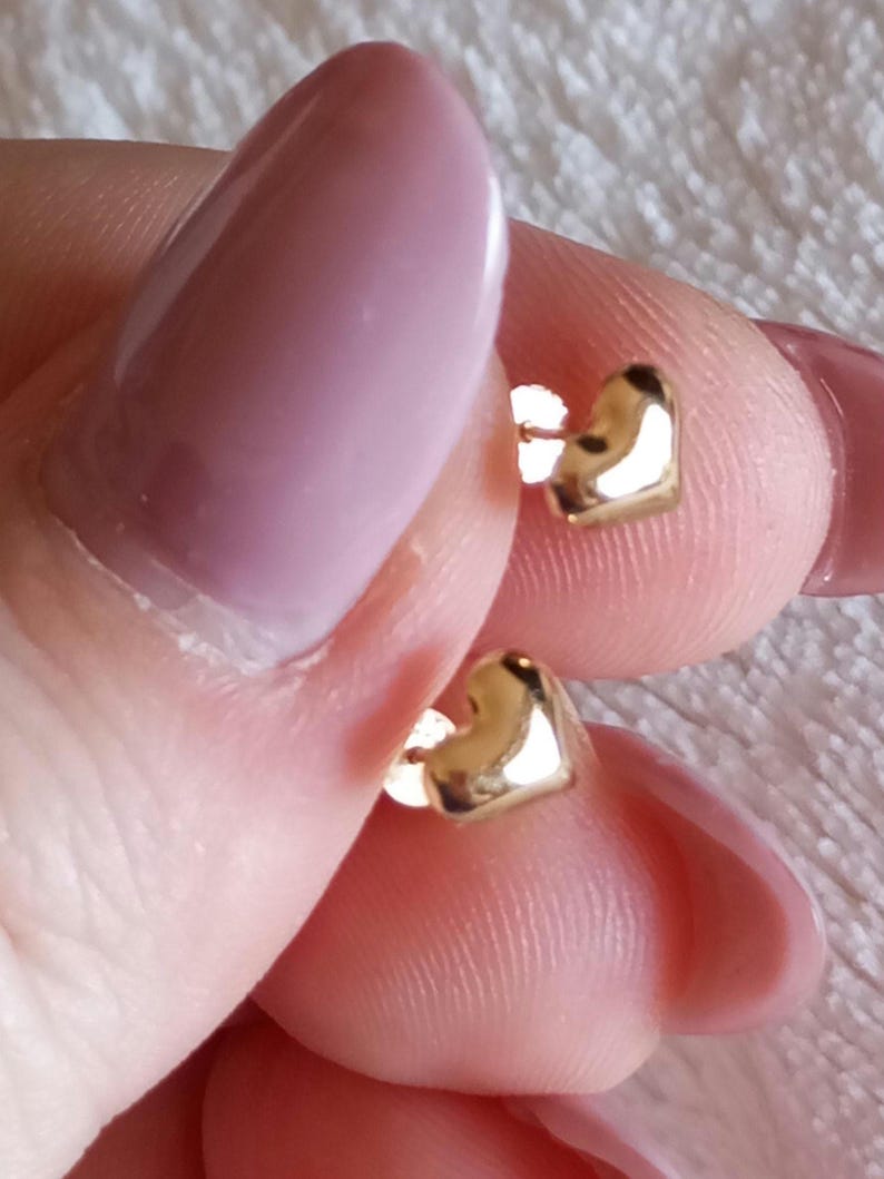 May include: A pair of small, polished gold-tone heart-shaped stud earrings. Each earring features a smooth, reflective surface in the shape of a classic heart, designed with a post back for secure attachment to the earlobe.