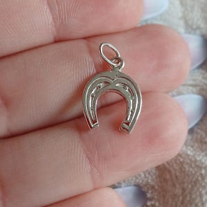 May include: A silver horseshoe pendant with a small loop at the top for a chain. The horseshoe has a curved shape with small decorative details. The pendant is from Minho Jewels.