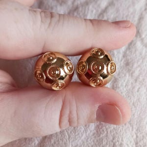 May include: A pair of gold-tone earrings with a round, textured design. The earrings are made of metal and have a shiny finish.
