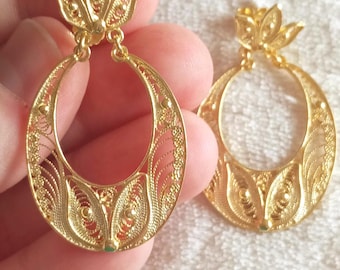 May include: A pair of gold-toned drop earrings featuring an intricate filigree design. Each earring consists of a circular hoop frame with delicate wirework patterns and a small, three-petaled floral motif attached to the top by a jump ring.