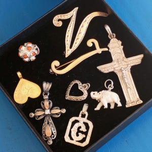 Peut inclure: Une collection de pendentifs et de breloques en argent et en or dans un coffret en velours noir. Les pendentifs comprennent un cœur, une croix, un éléphant, une lettre 'C' et une figure religieuse. Il y a aussi une broche en or en forme de lettre 'V' et une perle de corail rouge et orange.