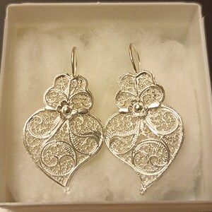 Pu&ograve; includere: Un paio di orecchini a forma di cuore in filigrana d'argento con un design floreale. Gli orecchini sono appesi a una scatola bianca.