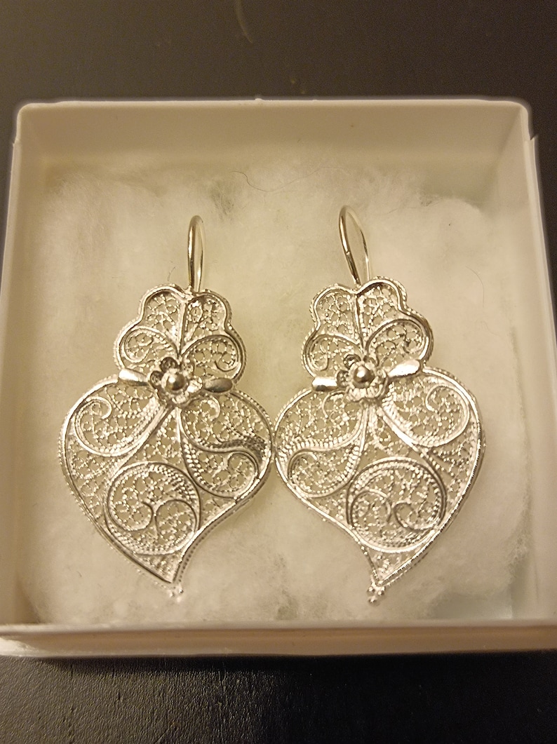 Pu&ograve; includere: Un paio di orecchini a forma di cuore in filigrana d'argento con un design floreale. Gli orecchini sono appesi a una scatola bianca.