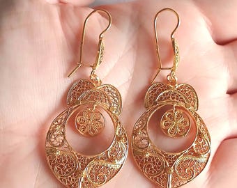 May include: A pair of gold-toned filigree drop earrings featuring an intricate openwork design. Each earring consists of a large circular frame with a smaller floral-shaped disc suspended in the center, topped with a curved hook wire.
