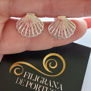 Può includere: Un paio di orecchini a lobo in argento a forma di conchiglie, caratterizzati da intricati motivi in filigrana. Sotto gli orecchini si trova un cartoncino nero con la scritta dorata FILIGRANA DE PORTUGAL e Minho Jewels in bianco.