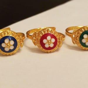 Puede incluir: Tres anillos dorados con bandas ajustables. Cada anillo presenta una pieza central circular esmaltada con un diseño de flor blanca. Las piezas centrales son de color azul, rojo y verde. Los anillos se exhiben sobre una superficie blanca.