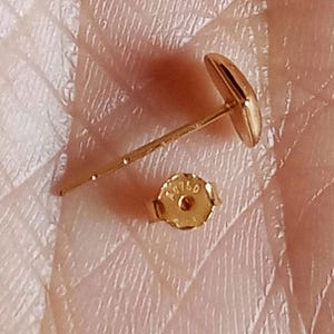 May include: A gold stud earring with a smooth, rounded face and a thin post. The accompanying gold earring back is circular with a scalloped edge and features the stamped text AU750, indicating the gold purity of the metal component.