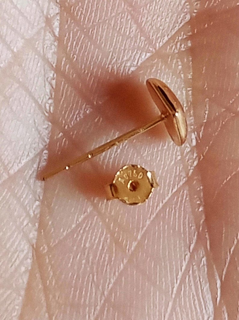 May include: A gold stud earring with a smooth, rounded face and a thin post. The accompanying gold earring back is circular with a scalloped edge and features the stamped text AU750, indicating the gold purity of the metal component.