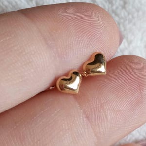 May include: A pair of small, polished gold-tone heart-shaped stud earrings. Each earring features a smooth, rounded, three-dimensional heart design with a reflective metallic surface, showcasing a simple and classic aesthetic for everyday wear.