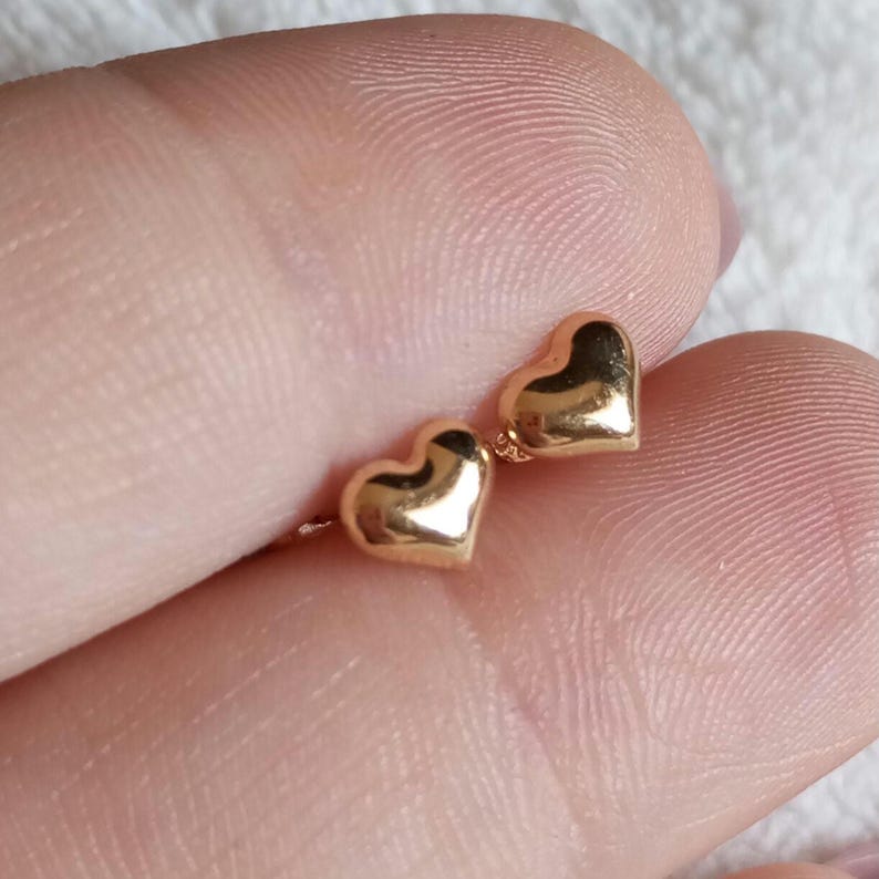 May include: A pair of small, polished gold-tone heart-shaped stud earrings. Each earring features a smooth, rounded, three-dimensional heart design with a reflective metallic surface, showcasing a simple and classic aesthetic for everyday wear.