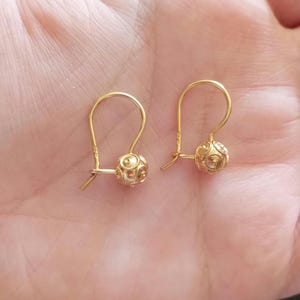 May include: A pair of gold earrings featuring small, spherical charms with intricate, embossed circular patterns. Each charm is suspended from a curved, hook-style wire closure, creating a delicate and polished appearance for these gold jewelry pieces.
