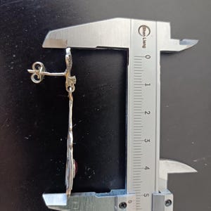 May include: A silver earring with a red gemstone and a decorative design. The earring is measured with a caliper, showing it is approximately 5.5 cm long.