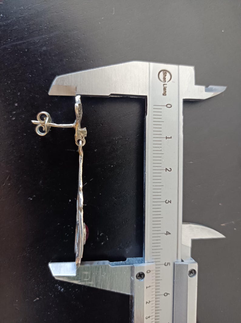 May include: A silver earring with a red gemstone and a decorative design. The earring is measured with a caliper, showing it is approximately 5.5 cm long.
