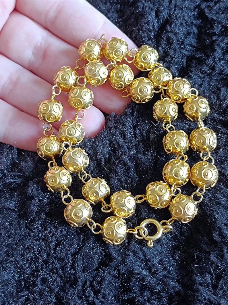 Pu&ograve; includere: Una collana d'oro con una catena e perline rotonde. Le perline hanno una superficie testurizzata con piccoli cerchi.