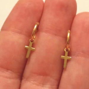 May include: A pair of gold-colored hoop earrings, each featuring a small cross pendant. The earrings are displayed against a blurred background, highlighting the jewelry's design and color.