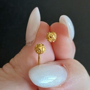 May include: A pair of small gold stud earrings featuring spherical heads with intricate filigree patterns and a central raised circular detail. The earrings have a polished gold finish and include a straight post for secure attachment to the earlobe.