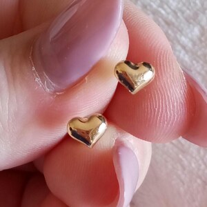 May include: A pair of small, polished gold-tone heart-shaped stud earrings. Each earring features a smooth, rounded, three-dimensional heart design with a reflective metallic surface. The earrings are shown as individual pieces held to display their shape.