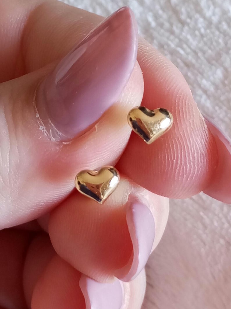 May include: A pair of small, polished gold-tone heart-shaped stud earrings. Each earring features a smooth, rounded, three-dimensional heart design with a reflective metallic surface. The earrings are shown as individual pieces held to display their shape.