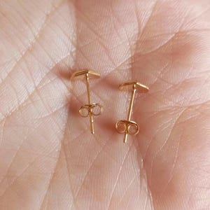 May include: A pair of small gold-toned stud earrings featuring a heart-shaped design. Each earring has a thin post and a secure butterfly-style back closure, showcasing a polished metallic finish that reflects light across the surface.
