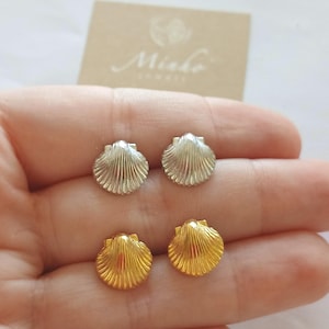 Könnte beinhalten: Zwei Paar muschelförmige Ohrstecker, ein Paar in Silberoptik und ein Paar in Goldoptik, mit vertikalen Rillen und fächerartigem Design. Die Ohrringe werden zusammen mit einer beigen Karte präsentiert, die den Schriftzug Minko trägt.