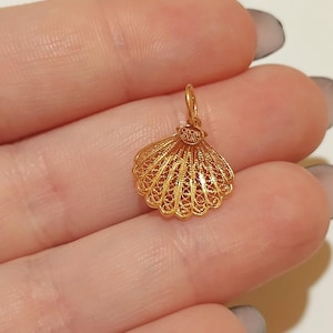 Könnte beinhalten: Ein goldener Muschel-Anhänger aus filigraner Arbeit mit aufwendigen Details. Der Anhänger hat eine kleine Schlaufe und den Text "Oficina Ouro". Der Anhänger wird in einer Hand gehalten. Der Text "Minho Jewels" befindet sich unten.