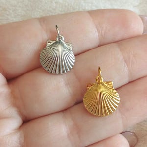 Op de afbeelding: Twee schelpbedels, één zilverkleurig en één goudkleurig, worden getoond. De bedels hebben een gedetailleerd, geribbeld ontwerp en een kleine lus om aan een ketting of armband te bevestigen. De bedels zijn klein, waarschijnlijk voor het maken van sieraden.