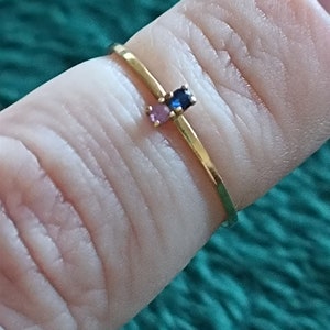 May include: A gold ring with three small gemstones: a blue sapphire, a pink sapphire, and a smaller pink sapphire. The ring is worn on a finger.