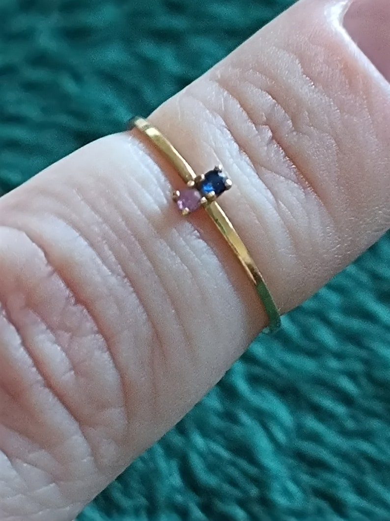 May include: A gold ring with three small gemstones: a blue sapphire, a pink sapphire, and a smaller pink sapphire. The ring is worn on a finger.