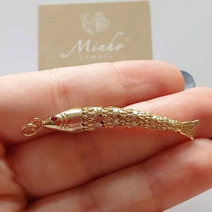 May include: A gold-toned articulated fish pendant featuring a textured, segmented body and a small red stone set as an eye. The fish has a smooth head section with etched gill details and a small circular jump ring attached to the mouth for hanging.