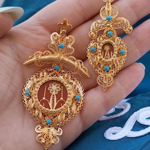 Könnte beinhalten: Aufwendige goldfarbene Filigranohrringe mit türkisfarbenen Akzenten. Jeder Ohrring hat ein zentrales Design mit einem rötlich-braunen Hintergrund und einem floralen Motiv. Die Ohrringe haben komplizierte, offene Muster, auf blauem Hintergrund.