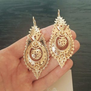 May include: A pair of gold-toned drop earrings featuring an intricate filigree design. Each earring has a large, elongated oval frame with a central spherical element, decorative cutouts, and a hook-style closure at the top for secure attachment.