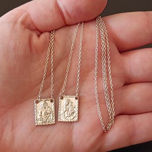 以下が含まれることがあります： 2つの金色のロザリオネックレス。各ネックレスは、エンボス加工された宗教的な人物が描かれた長方形のペンダントが特徴です。ペンダントは繊細なチェーンに接続されています。ネックレスは手の上に表示されています。