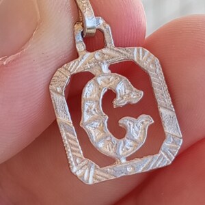 May include: Silver charm pendant featuring the letter G in a decorative, ornate frame. The letter is cut out and the frame is detailed with intricate lines and patterns.