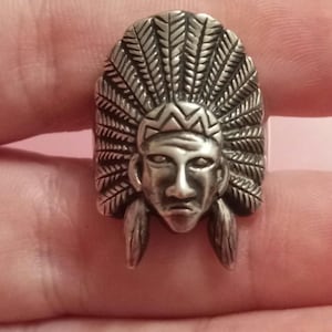 Op de afbeelding: Een zilverkleurige ring met een gedetailleerd ontwerp van een Indiaanse hoofdtooi. De ring toont een gezicht met gedefinieerde kenmerken en veerdetaillering, wat een opvallend en cultureel sieraad creëert.