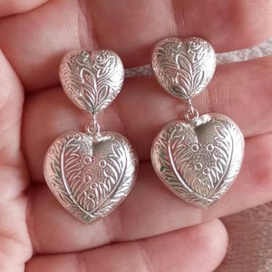 May include: A pair of silver heart-shaped dangle earrings with a detailed floral design. The earrings are made of metal and have a polished finish.