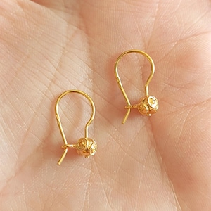 May include: A pair of gold earrings featuring small, spherical charms with intricate, textured patterns. Each earring is suspended from a curved, hook-style wire, showcasing a polished, reflective finish that catches the light.
