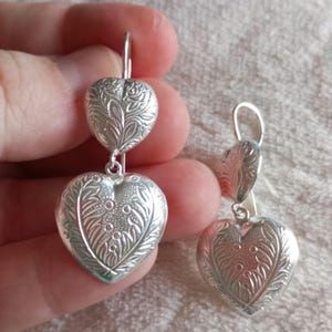 May include: Silver dangle earrings with two heart-shaped charms. Each charm has an intricate floral design.
