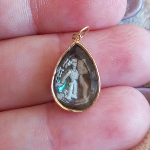May include: A teardrop-shaped pendant with a gold-colored frame. The pendant features a detailed, miniature carving of a figure in a classical pose. The carving is set against a dark background, and the pendant has a small loop at the top for a chain.