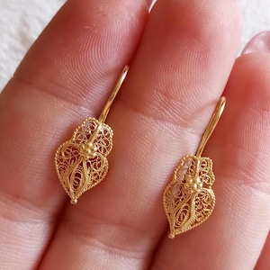 Könnte beinhalten: Ein Paar goldene Filigranohrringe. Jeder Ohrring hat ein herzförmiges Design mit aufwendigen Durchbrucharbeiten und einem kleinen floralen Akzent. Die Ohrringe haben ein zartes, verziertes Aussehen.