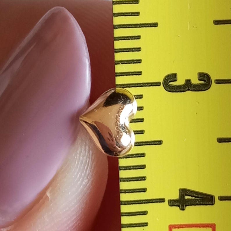 May include: A small, polished gold-tone heart-shaped charm positioned next to a yellow measuring tape. The heart measures approximately 0.25 inches in width, as indicated by the markings on the visible ruler scale next to the object.