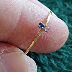 May include: A delicate gold ring with a small blue and pink gemstone accent. The ring is thin and simple, with a minimalist design.