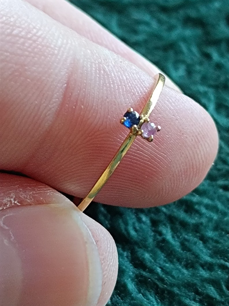 May include: A delicate gold ring with a small blue and pink gemstone accent. The ring is thin and simple, with a minimalist design.