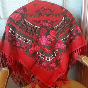 May include: A vibrant red shawl with a floral pattern featuring pink and red roses. The shawl has a fringed edge and is draped over a chair. The design includes green and brown decorative elements.