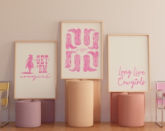May include: Three framed prints with pink text and illustrations on a white background. The prints are on a shelf with pink and beige cylindrical stands. The left print says "Get 'Em Cowgirl" and features a pink silhouette of a cowgirl riding a horse. The middle print features a pink illustration of cowboy boots. The right print says "Long Live Cowgirls" with a pink heart.