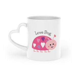 Może przedstawiać: Biały kubek ceramiczny z uchwytem w kształcie serca. Kubek ma różowego biedronkę z fioletowymi i białymi sercami na plecach i nadrukiem "Love Bug" z przodu.