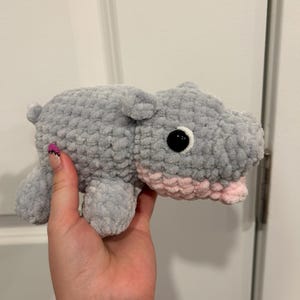 May include: A handmade, gray crochet hippo plush toy with a pink mouth and black eyes. The plush animal is soft and cuddly, perfect for a child's toy collection or nursery decor.