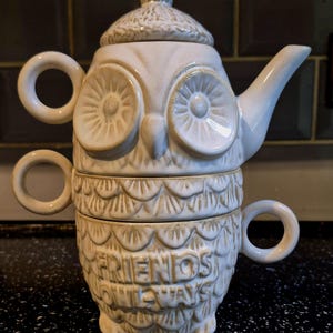 May include: A cream-coloured owl-shaped teapot set. The teapot has a lid and a spout, with two stacked teacups below. The cups and teapot are decorated with an owl face and feather-like patterns. The words "FRIENDS OWLWAYS" are embossed on the bottom cup.