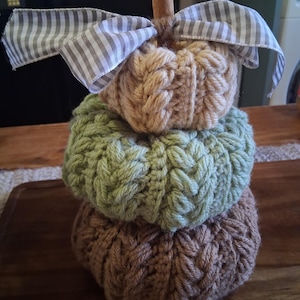 May include: A stack of three knitted pumpkins in shades of brown, green, and tan, topped with a cinnamon stick stem and a gray and white striped bow. The pumpkins are arranged on a wooden surface, creating a fall-themed decoration.