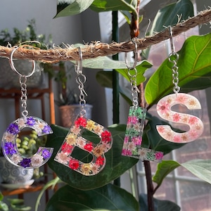 Op de afbeelding: Vier hars sleutelhangers met de letters "CBSL" hangen aan een touw. Elke letter is gevuld met kleurrijke gedroogde bloemen. De letters zijn bevestigd aan zilveren sleutelringen.