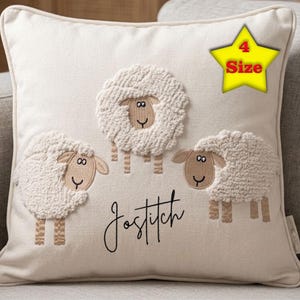Op de afbeelding: Crèmekleurig vierkant kussen met drie geborduurde schapen met pluizige witte wol en bruine poten. Het woord "Jostitch" staat eronder in een sierlijk lettertype. Een gele ster met het woord "Size" staat rechtsboven.